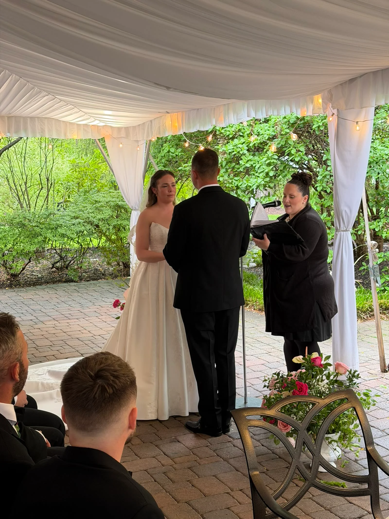 Guests watch an outdoor wedding ceremony in a garden setting.